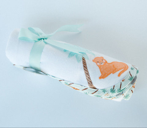 A swaddle blanket with a white background featuring an orange and green applique in the shape of a leaf and a blue and white striped ribbon.