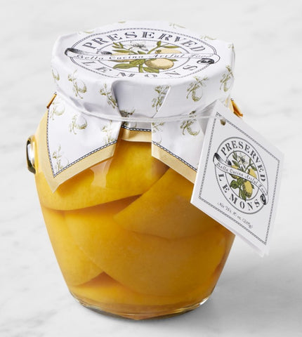 A glass jar of Preserved Lemons filled with sliced yellow lemons, sealed with a white patterned lid and ribbon, with a hanging tag on a light marble surface.