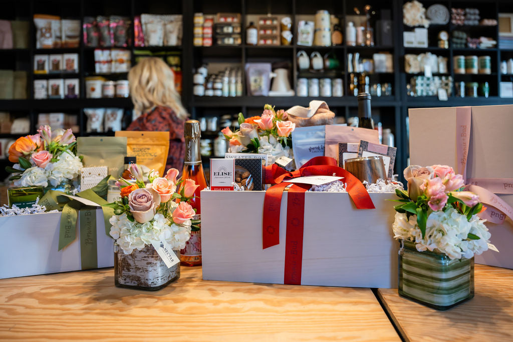 A gift basket arrangement on a wooden table in a gift shop, featuring a large white box with a red ribbon and smaller floral arrangements, with shelves of products blurred in the background.