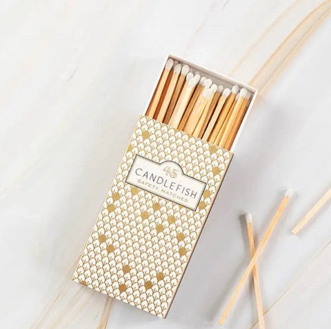 A box of Candlefish matchsticks with gold foil geometric patterns on a white marble background, with a few loose matchsticks scattered nearby.