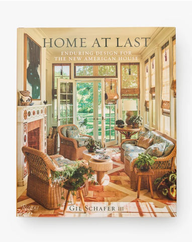 The book cover of 'Home At Last: Enduring Design For The New American House' by Gil Schafer III, featuring a warm, sunlit interior room with wicker furniture, a fireplace, and large glass doors.