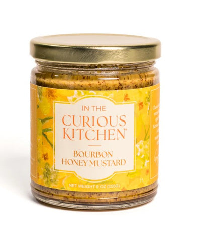A glass jar of In the Curious Kitchen Bourbon Honey Mustard with a gold metal lid and a yellow label, set against a plain white background.