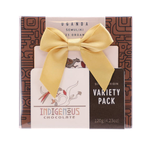 A square gift box of Indigenous Chocolate Variety Pack from Uganda, decorated with a large yellow satin bow. The packaging features a brown geometric pattern on the sides, a cream-colored center, and a black label on the right side that reads 'VARIETY PACK'.