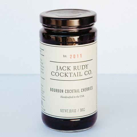 A jar of Jack Rudy Cocktail Co. Bourbon Cocktail Cherries with a white label, showcasing the brand and product name.