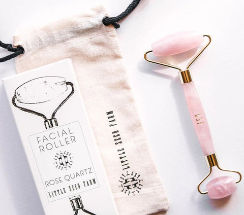 A pink rose quartz dual-ended facial roller tool with gold-tone metal accents, shown next to its beige drawstring pouch and a white product box labeled 'Facial Roller Rose Quartz Little Seed Farm'.