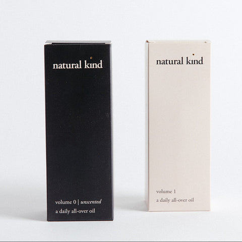 Two cylindrical bottles of Natural Kind All-Over Oil, one standing upright and the other lying down, both with black and beige labeling.
