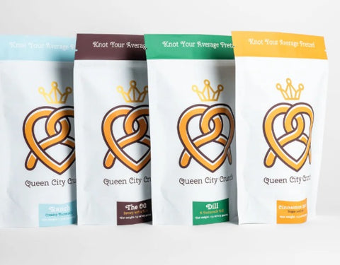 Four stand-up pouches of Queen City Crackerbread snacks in different flavor variants, lined up side-by-side on a white background. The packages are white with a prominent orange heart-shaped pretzel logo and a gold crown graphic.