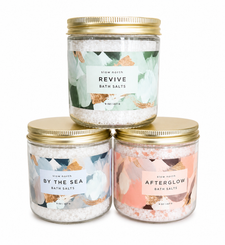 Three glass jars of Slow North bath salts stacked in a pyramid on a white background. The top jar is labeled 'REVIVE BATH SALTS' with a green and white floral label and a gold lid.