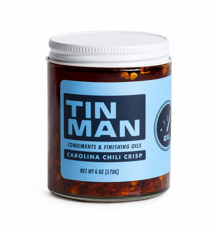 A glass jar of Tin Man Carolina Chili Crisp with a white lid and a light blue label, set against a plain white background.