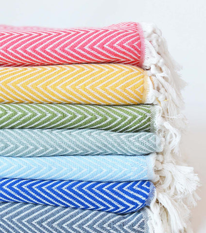 A stack of folded woven towels with white fringe, shown in multiple colors including pink, yellow, green, light blue, royal blue, and gray. Each towel features a white herringbone pattern on a colored background.
