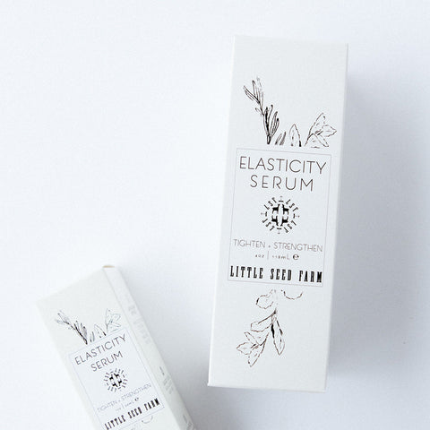 A white bottle with a label that reads 'ELASTICITY SERUM' from Little Seed Farm, placed on a white surface.
