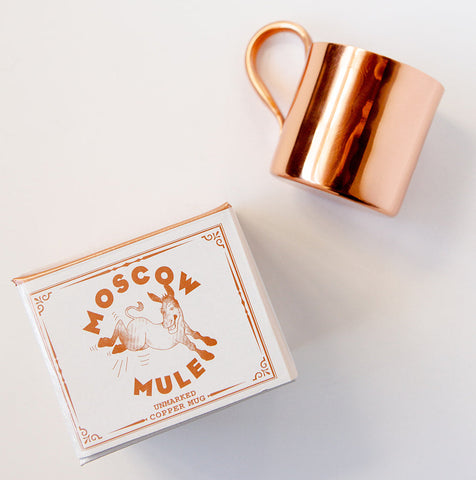 A copper mug with a matching lid, placed next to a white box with 'Moscow Mule' written on it.